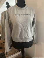 Gray Club Monaco sweatshirt with 'CLUB MONACO' printed text on chest, displayed on mannequin in front of mirror.