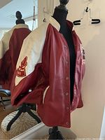 Front side of the jacket on a dress form, showing red leather body with white sleeves and collar, snap button closure, and embroidered patch on left chest.