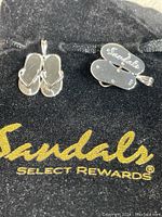 Two sterling silver flip-flop sandal charms on black Sandals Select Rewards pouch, showing front and back of charms