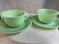 Two light green jadeite cups set on matching saucers viewed from front.