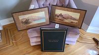 Two framed landscape art prints and one hardcover book displayed on a chair and the floor