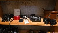 Shelf view showing all items: Birks binoculars and case at left and right, compact digital camera and charger in center, black Sport film camera and small binoculars at right.