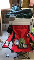 Red camping chair with several camping items placed on and around it including a boxed portable stove and accessories on the seat.