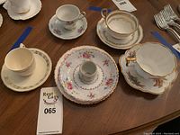 Photo showing several china tea cups and saucers arranged in a group on a wooden table, variety of floral designs and gold trim visible.