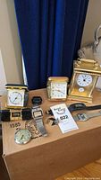 Full view of all clock and watch items on a cardboard box: Damas pocket watch, carriage clock, multiple wrist watches with various straps and face styles