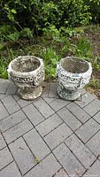 Photo showing two weathered concrete garden planters with Greek key decorative bands and pedestal bases on a paved outdoor surface near greenery.
