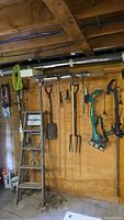 Overall view of assorted garden tools including hand tools, step ladder, and power tool hanging on wooden wall