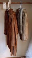 Two fur garments hanging on hangers: a long brown E. Anderson fur coat and a short Hudson's Bay fur jacket with a separate gray fur neck piece.