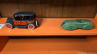 Two pressed tin toy cars displayed on an orange shelf against a wood-paneled wall: one vintage-style orange and black car on the left, a green streamlined race car on the right.