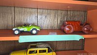 Shelf displaying three pressed tin toy vehicles: green sports car, red tractor, and yellow sedan toy car partially visible below