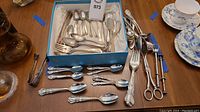 Full assortment of silver plated utensils including forks, spoons, and specialty tools all arranged on a wooden surface and some in a blue box.