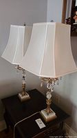 Pair of brass table lamps with white rectangular fabric shades on a dark table near a wall.