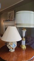 Two table lamps on a wooden table, one geometric marble base with beige shade, the other taller white ceramic and brass lamp with Greek key trim shade
