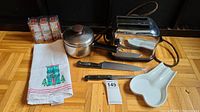 Overview photo of entire lot on floor including toaster, knives, saucepan, towel, ceramic dish, and spice tins.