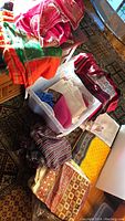 Photo showing multiple stacks of colorful silk and fine fabrics including orange, green, red, white, and patterned pieces in a plastic tote on carpet