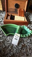 Wooden humidor open showing cigar holder, humidity gauge, instruction sheet, small black container, and plastic bottle with red cap nearby. Green ceramic ashtray placed in front.