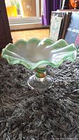Murano art glass footed fruit bowl with scalloped rim, green edge and swirl pattern on stem, shown on carpet with window light.