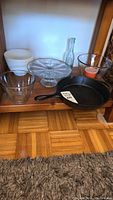 Cast iron skillet combined with glass mixing bowls, cake stand, milk glass items on shelf with visible wooden floor and carpet.