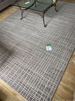 Full view of the rectangular grey, beige, and cream area rug with grid pattern under a glass coffee table and sofa on wood flooring.