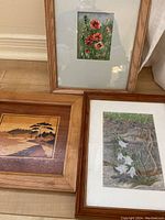 Three framed art pieces on floor, including two floral paintings and one wood inlay landscape art