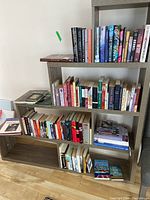 Books arranged on a wooden shelf with spines showing various titles and authors, all subject to wear and usage.