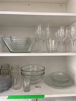 Clear glass bowls, wine glasses, tumblers, and plates arranged on a shelf, showing assortment and size variety