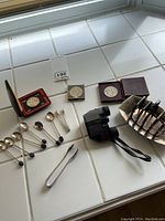 Full view of lot items arranged on tile surface including coins in display case, silver spoons, toast rack, tongs, and binoculars.