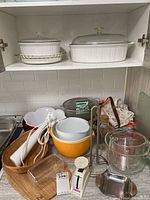 Full shelf showing assortment of CorningWare casserole dishes, glass bakeware, measuring cups, mixing bowls, white kitchen utensils, a glass cutting board, aprons, and tea towel.