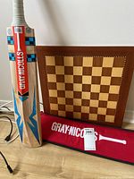 Full view of the cricket bat, red protective cover, and wooden checkerboard game board on floor against wall.