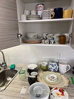 Photo of upper shelf showing stacked plates, mugs of various patterns and colors including black and white geometric print and solid yellow mug, along with containers and bowls.