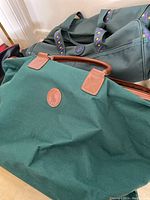 Two green duffel bags resting on the floor, one lighter with brown leather handles and one darker with circular leather patch and handles visible