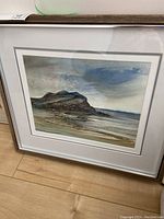 Framed watercolor artwork showing coastal rock formation and tidal landscape under a cloudy sky, signed Mack Sloan.