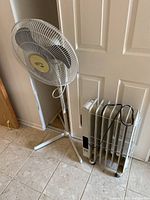 Wide shot showing Hampton Bay white floor fan and DeLonghi portable oil-filled heater side by side near a door with tiled floor.
