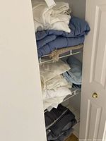 Shelf with multiple folded blankets and sheets in cream, white, blue, and gray tones.