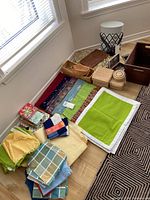 Photo showing assortment of baskets, woven trays, and linen items laid out on floor near a window, illustrating range and condition.
