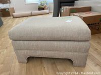 Front view of the beige upholstered ottoman showing fabric texture and base design.