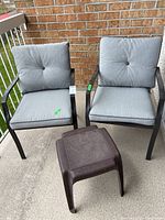 Two black metal patio chairs with grey cushions and a brown small plastic side table arranged on a balcony.