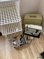 Vintage White model 710 sewing machine with power cord and foot pedal beside beige carrying case. Three white plastic stacking bins and box of assorted sewing accessories on floor.