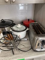 Overview of various small kitchen appliances on countertop including Crockpot, rice cooker, 4-slice toaster, red fondue pot, and coffee grinder with cords and forks visible.