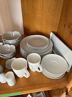 Overview of stacked white china including various sized plates, bowls with scalloped edges, mugs, and salt and pepper shakers.