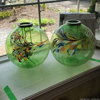 Two round hand blown green glass candle holders with a multicolored leaf design on a windowsill.