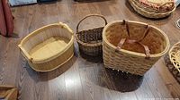 Three wood and woven baskets placed on a wooden floor. Includes a small dark brown wicker basket, a large light wood woven basket with brown handles, and an oval light wood basket.