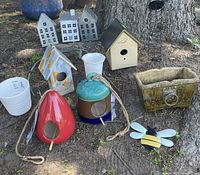 Selection of bird feeders and candle holders arranged outdoors on the ground by a tree trunk, showcasing variety of styles and materials