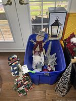 Photo showing all the Christmas decorations including the wood snowman, ceramic lighted trees, ceramic Happy Holidays pitcher, decorative star, lantern in box, and blue storage bin with other items inside.
