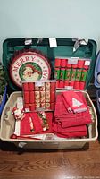 Photo of holiday items stored in a beige plastic bin with green lid, showing metal Santa Merry Christmas pop lid, two boxes of Christmas crackers, and stacked towels and linens.