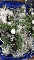 Blue plastic bin filled with mixed silver and white Christmas decor items including flocked greenery with ornaments, glittered garland, and silver reindeer figurines.