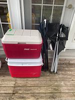 Two hard-sided coolers stacked vertically with two folded camp chairs leaning against them on a wooden deck outdoors.