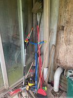 Full view of various garden tools including broom, axe, shovel, shears, and handles leaning against a wall.