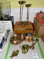 Overview of all items showing copper kettles, brass candle holders, vases, ladle, and fish ashtray arranged on orange tray with boxes and wall background.