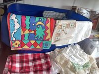 Blue plastic bin containing various linens, colorful printed linen, white embroidered linen, red checked fabric, and lace doilies on table surface
